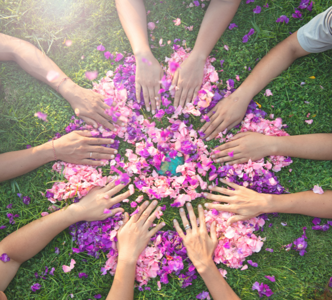 Mehrere Hände legen bunte Blütenblätter zu einem Kreis auf grünem Gras